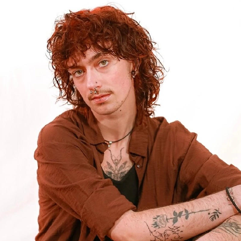 A young person with curly, shoulder-length brown hair and facial piercings sits against a white background. They wear a brown shirt over a black top, have visible tattoos on their chest and forearm, and look directly at the camera with a calm expression.