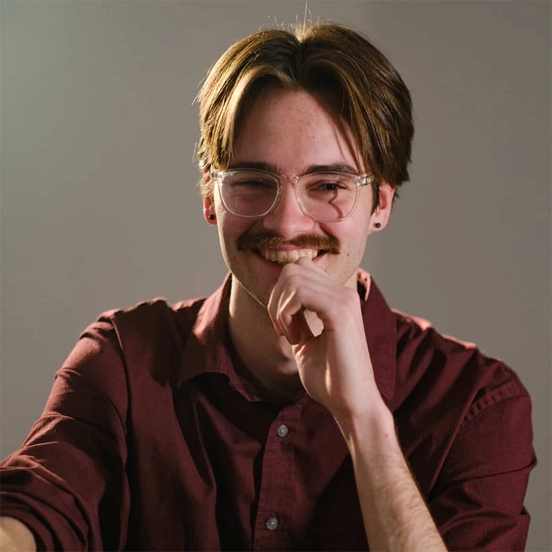 A young person with light skin, brown hair, a mustache, and glasses smiles warmly while wearing a maroon button-up shirt. They rest one hand near their face and sit against a plain, neutral background. The lighting is soft, creating a friendly atmosphere.