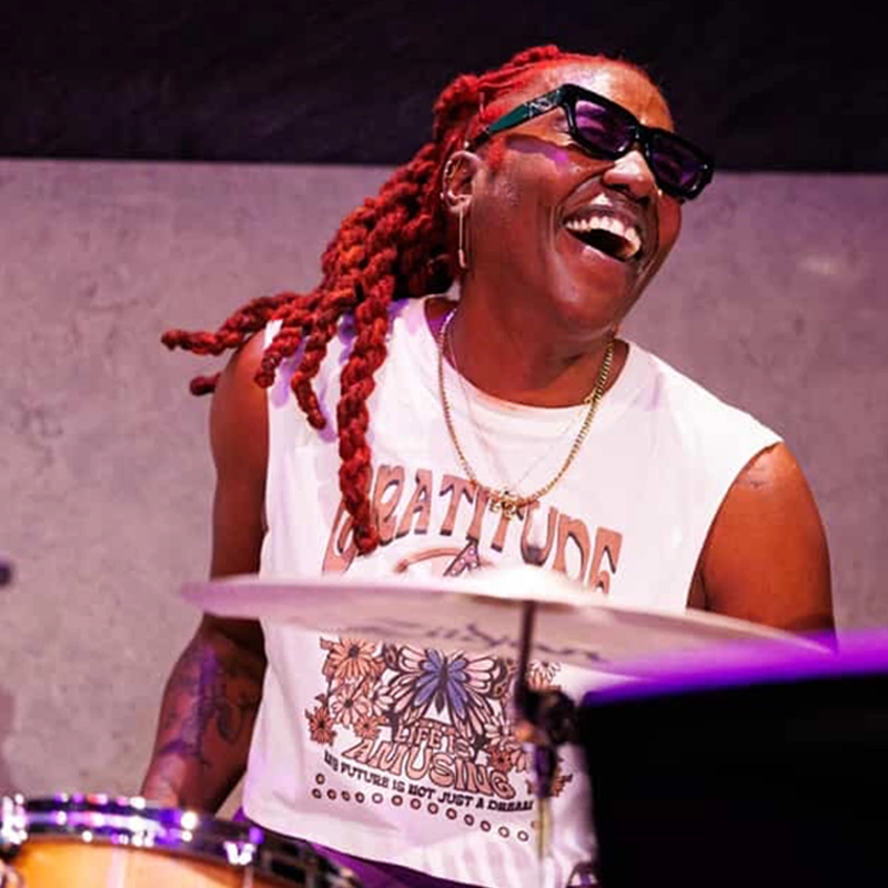A smiling person with bright red dreadlocks, sunglasses, and a sleeveless 