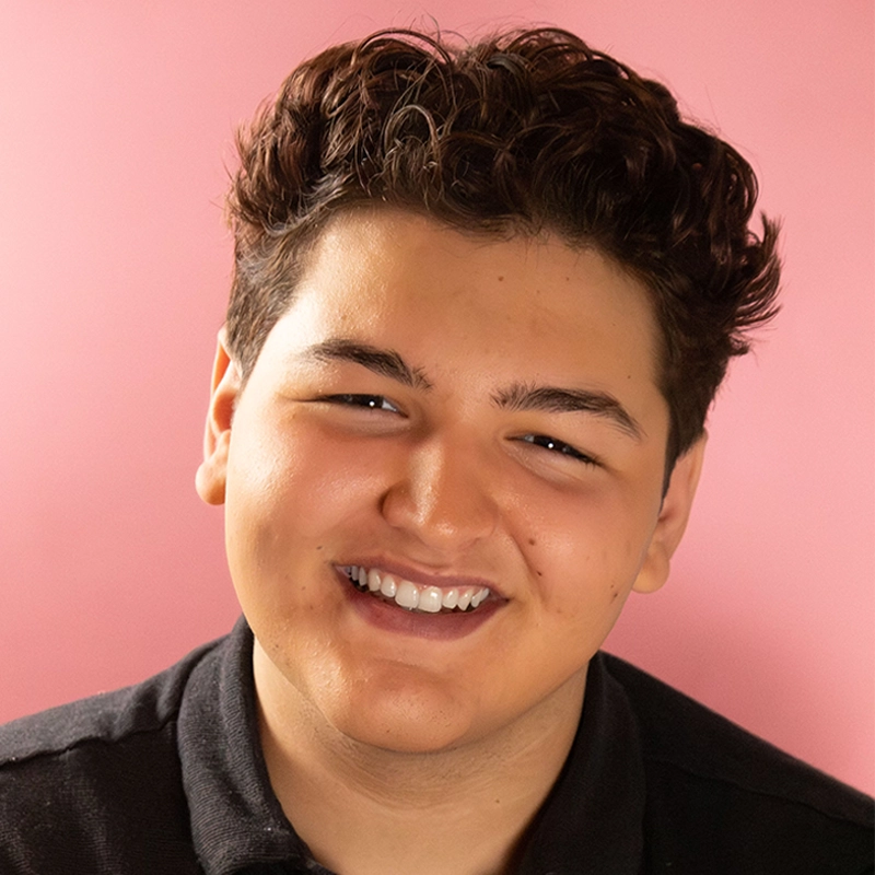 Jacob Phillips, a young person with short, wavy brown hair, smiles warmly at the camera. They have light skin and wear a black collared shirt. The soft pink background creates a pleasant and friendly atmosphere.