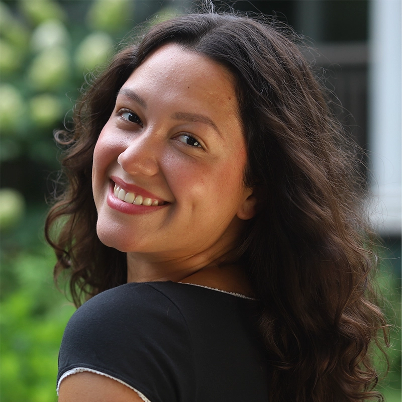 A woman with long, wavy brown hair smiles over her shoulder at the camera. She wears a black top with white trim and stands outdoors with a blurred background of greenery and flowers. The lighting is natural, giving her face a warm, vibrant look.
