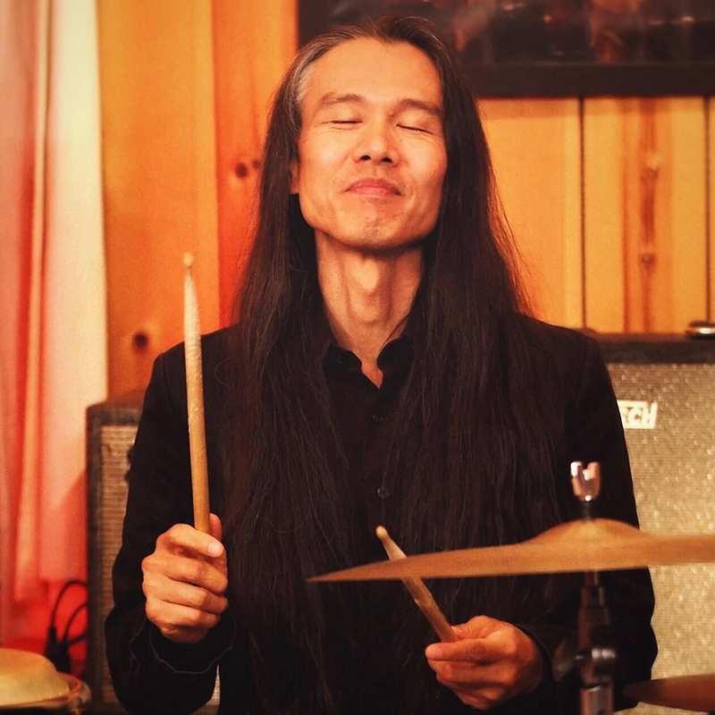 A man with long black hair and a serene smile plays drums with his eyes closed. Kendal Hartse, dressed in a black shirt, holds drumsticks and appears absorbed in the music beside wooden paneling and sound equipment in a warm, indoor setting.