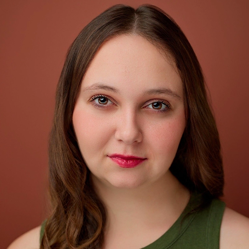 A young woman with fair skin, long brown hair, and blue eyes looks directly at the camera. She has a slight smile and wears a green sleeveless top. The background is a solid warm brown color. Her makeup is natural with pink lipstick, highlighting the effortless elegance of Isabella Concetta Borte.