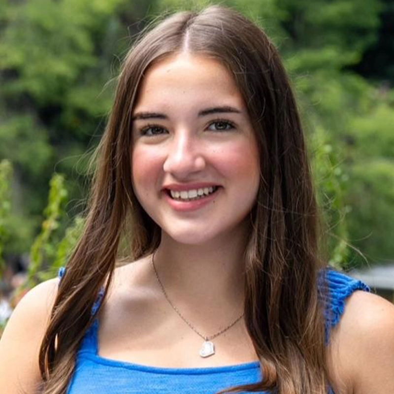 Isabella Fortunato, a young woman with long brown hair and fair skin, smiles at the camera. She wears a blue sleeveless top and a silver heart-shaped pendant, with blurred green trees in the background suggesting an outdoor setting.