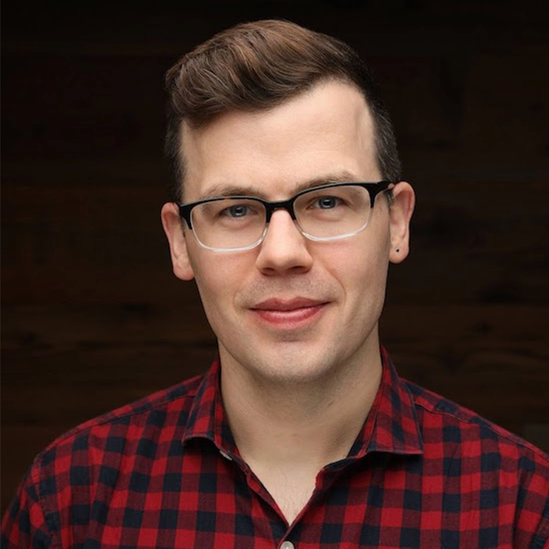 A man with short brown hair and glasses, wearing a red and black plaid shirt, smiles gently at the camera. The background is dark and out of focus, highlighting his face and upper torso.