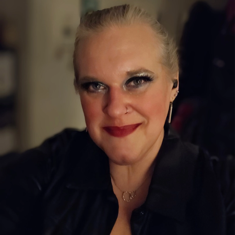 A smiling woman, Julia Schade, with blonde hair pulled back, bold eye makeup, red lipstick, and a black top. She has a nose ring, earrings, and a necklace, posing against a softly blurred indoor background.