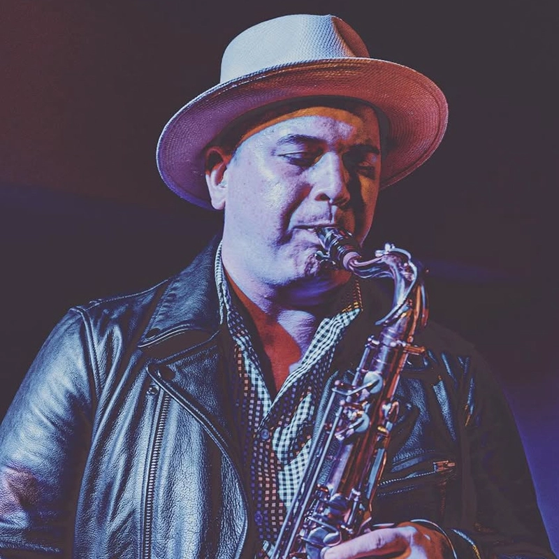 A person wearing a light-colored fedora and black leather jacket plays a saxophone. The background is dark, and stage lighting casts blue and purple tones on their face and clothing. The individual appears focused on performing music.