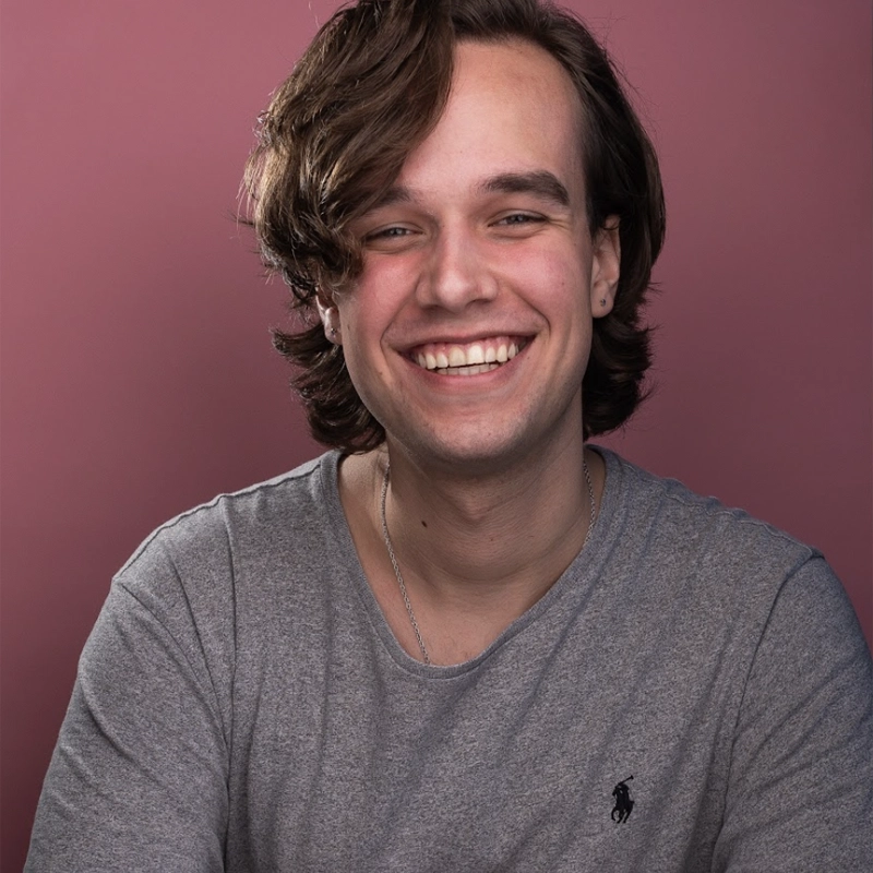 Young man with wavy brown hair and a broad smile poses against a pink background. He wears a grey T-shirt with a small dark logo on the chest. His posture is relaxed, and he appears happy and approachable, with soft lighting highlighting his face.