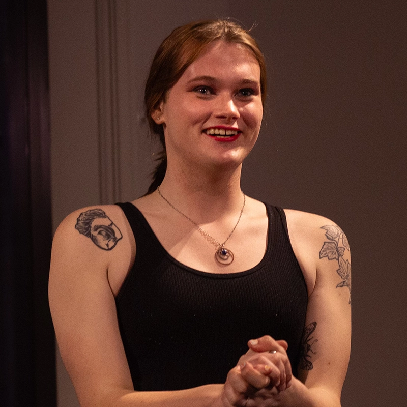 A smiling person with fair skin, brown hair tied back, and red lipstick stands indoors, clasping their hands. They wear a black sleeveless top, a circular pendant necklace, and have tattoos on both shoulders—one of a face and one of maple leaves.