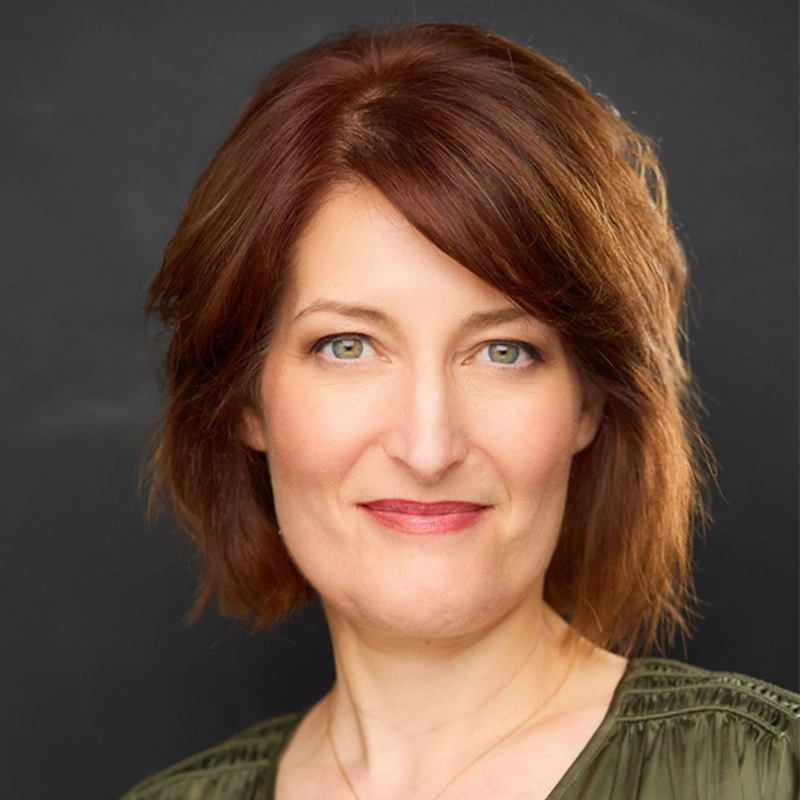 A woman with fair skin and shoulder-length reddish-brown hair smiles softly at the camera. She wears a green top and a subtle necklace. The dark, plain background draws focus to her face, while even lighting highlights her features—Sawyer Whitted.