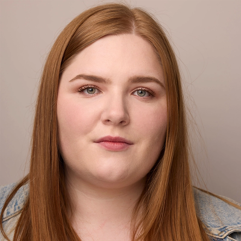 A young woman with straight, long reddish-brown hair and fair skin looks directly at the camera. She has light makeup, full lips, and faintly arched eyebrows. Styled like Sawyer Whitted, she wears a light denim jacket against a plain beige background.
