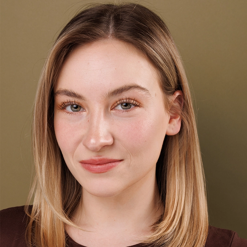 A young woman with straight, shoulder-length blonde hair and fair skin is looking at the camera with a slight smile. She has light-colored eyes, natural makeup, and is wearing a brown top. The background is plain olive green.
