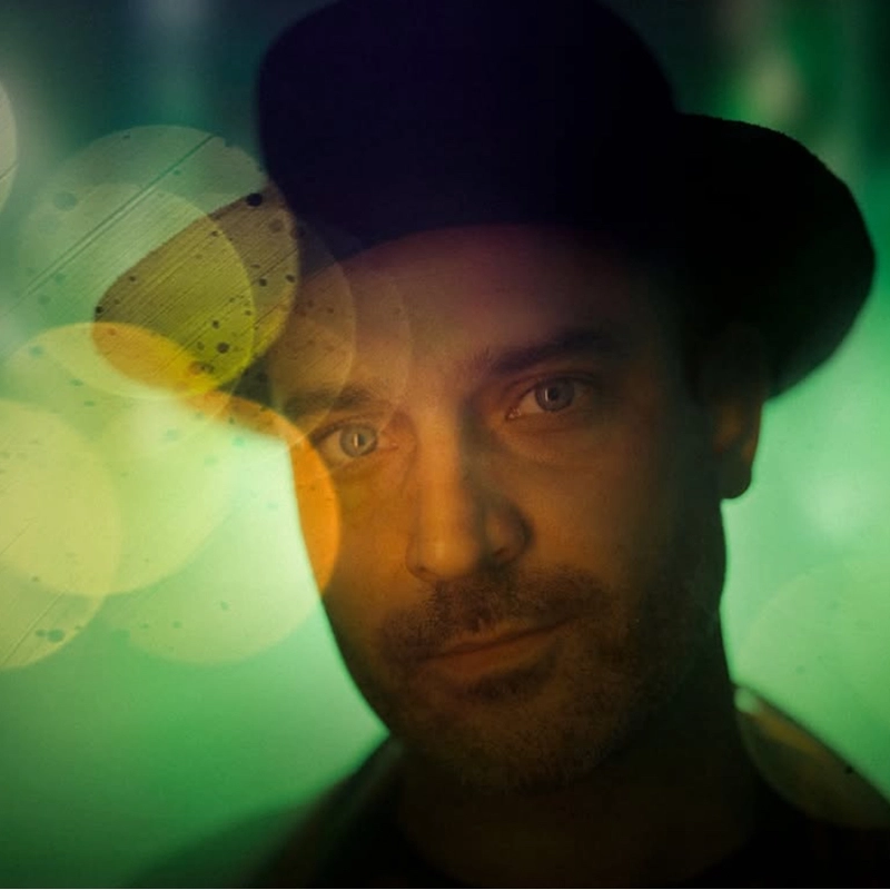 A man wearing a black hat gazes softly at the camera. The image has a dreamy, artistic effect with greenish lighting and circular, golden bokeh lights overlapping his face, creating a surreal and atmospheric mood.