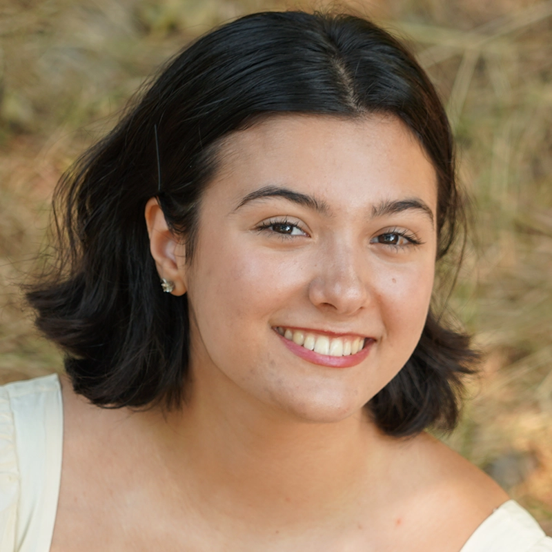 A young woman with short, dark hair and light skin smiles warmly at the camera. She wears a cream-colored top and small earrings. The soft, out-of-focus grass and earthy tones behind her evoke the natural charm of a Sawyer Whitted portrait.