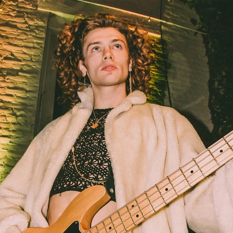 A person with curly hair, wearing a fluffy cream-colored coat and a black lacy crop top, plays an electric bass guitar. Warm lighting and a brick wall background create an intimate, lively atmosphere. The person looks upward, appearing focused and expressive.