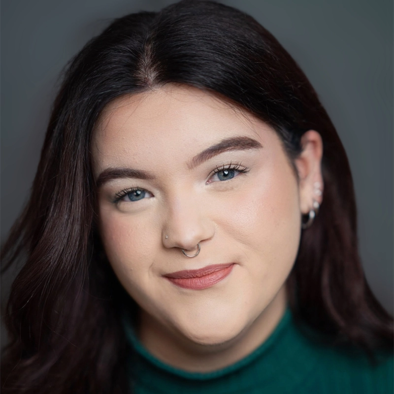 A young woman with fair skin and long dark brown hair smiles softly. Reese Maynard has light blue eyes, subtle makeup, a nose ring, and multiple earrings. She wears a green top against a neutral gray background with soft focus.