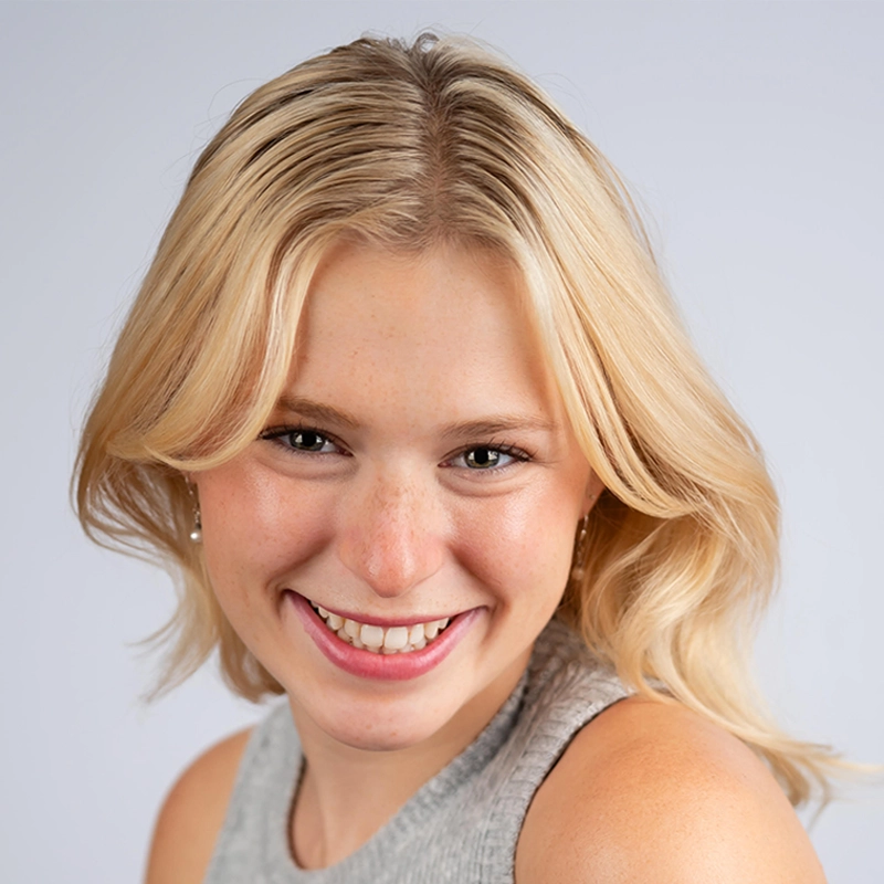 Isabella Fortunato, a young woman with long blonde hair, smiles warmly at the camera. She has light skin, is wearing a sleeveless gray top, and is posed against a plain light gray background. Her hair is parted in the middle and softly frames her face.