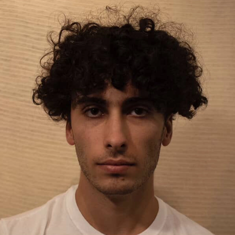 A young man with curly dark hair and a short beard looks directly at the camera with a neutral expression. He is wearing a plain white shirt and stands in front of a beige, textured background. The lighting is soft and warm.