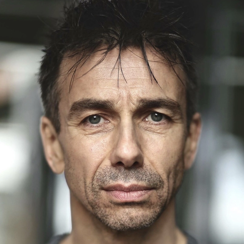 A middle-aged man, resembling Jon Peterson, with short, dark, messy hair and blue eyes stares directly at the camera with a neutral expression. He has light skin, some facial stubble, and is wearing a gray shirt. The background is blurred and dark.