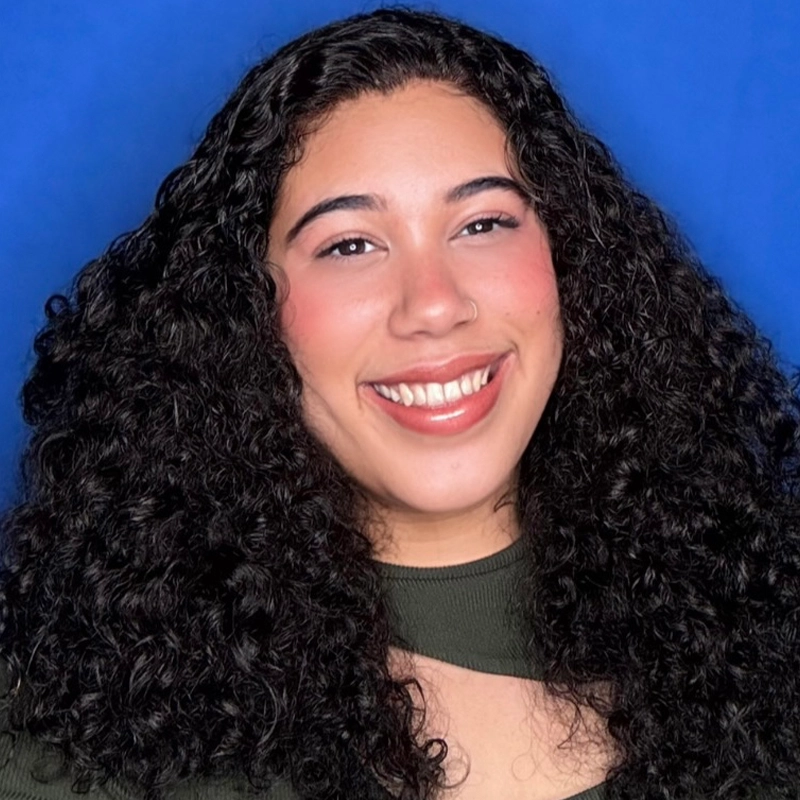 A young woman with long, dark, curly hair smiles warmly. She has medium skin, pink blush, natural makeup, and glossy lips. She wears a dark green top with a cutout at the neckline. The background is a solid bright blue.