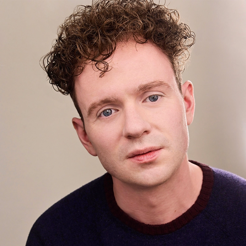 A young man with curly brown hair and fair skin, Sawyer Whitted wears a dark sweater with a maroon collar, looking softly at the camera. The plain, light background emphasizes his facial features and gentle expression.