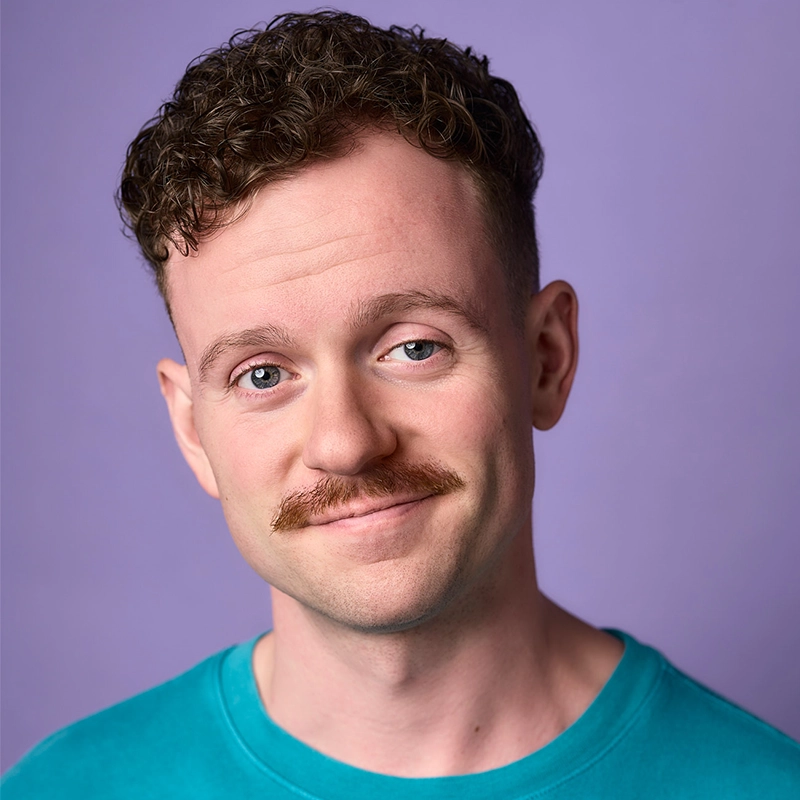 Sawyer Whitted, a young man with short curly brown hair and a light brown mustache, smiles gently. He wears a teal shirt and stands against a plain lavender background, head slightly tilted as he looks directly at the camera.