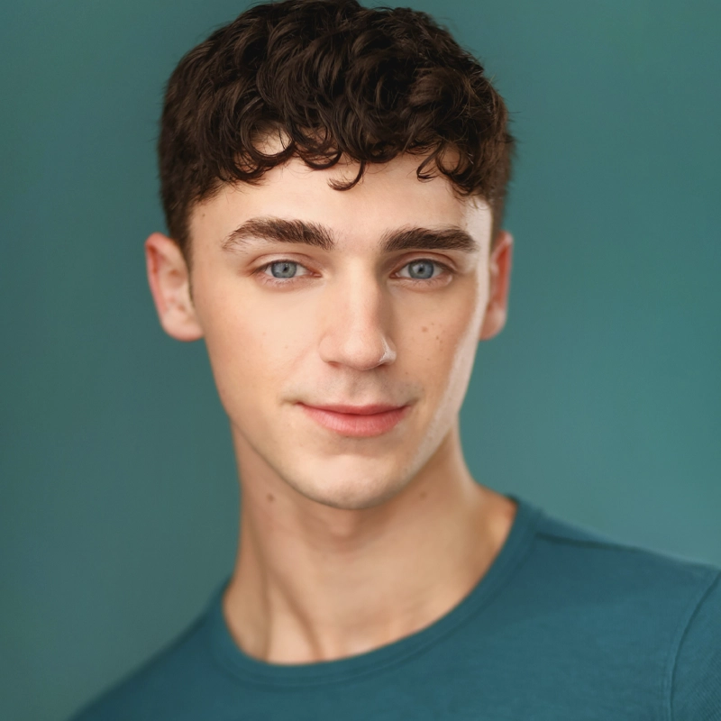 A young man, Jake Siffert, with short, curly dark brown hair and blue eyes is smiling slightly. He is wearing a teal shirt and posed in front of a solid teal background. The soft lighting highlights his clear skin and facial features.