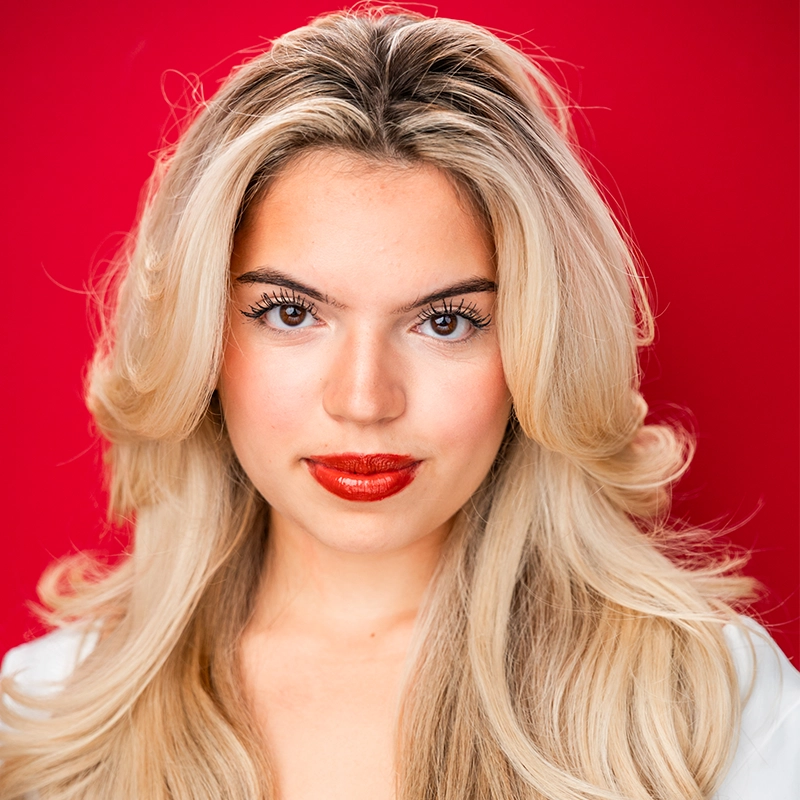 Isabella Fortunato, with long, wavy blonde hair and bold red lipstick, poses against a vibrant red background. Her defined eyebrows, long eyelashes, and confident gaze pair perfectly with her light-colored top and neutral expression.