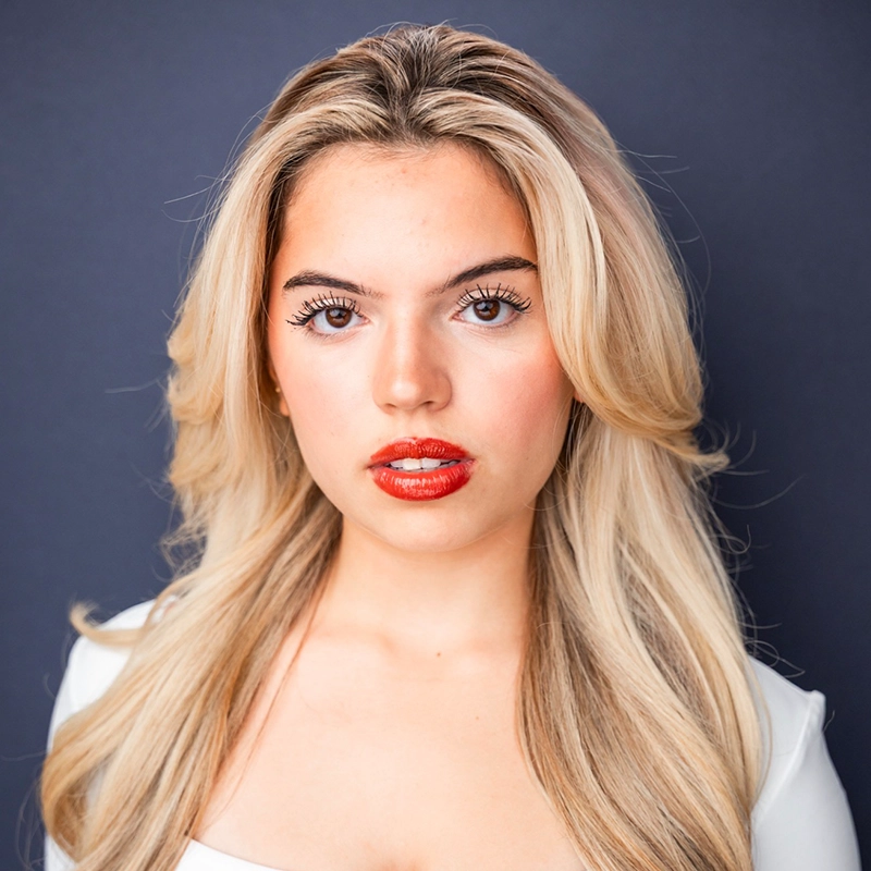 A young woman, Sofia Thérèse Maroulis, with long, wavy, platinum blonde hair poses in front of a dark gray background. She wears a white top and bold red lipstick, defined eyelashes, and minimal makeup, looking directly at the camera with a neutral expression.