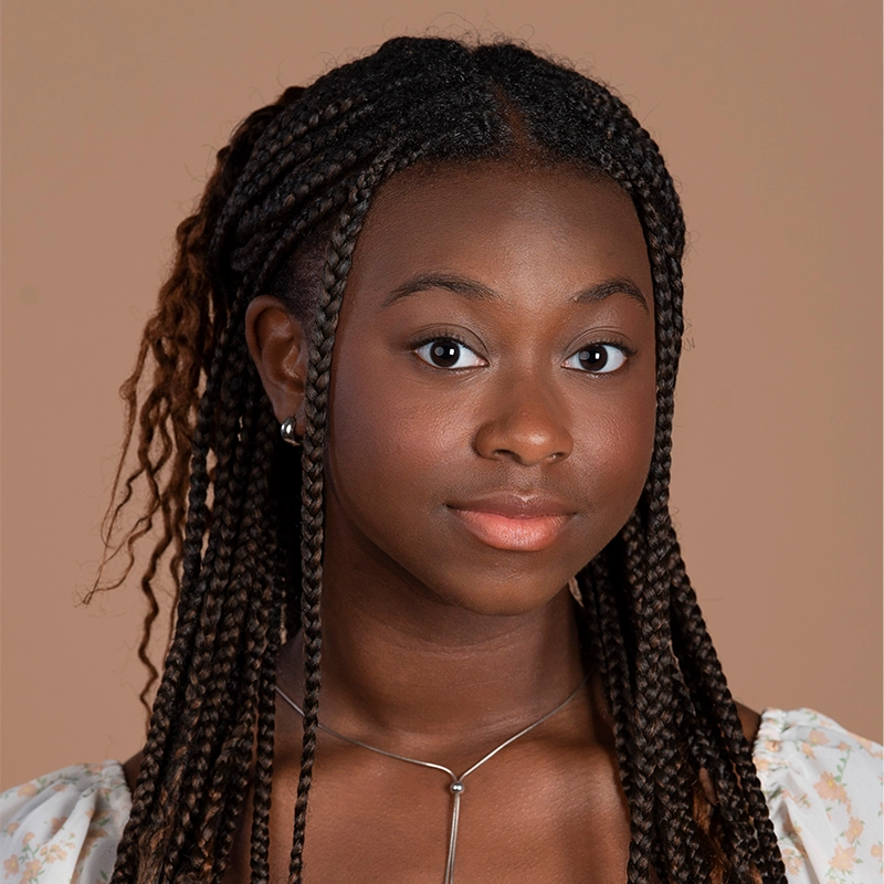 A young woman with long, dark braided hair poses against a beige background. She wears a light floral top, a silver necklace, and small stud earrings. Her expression is calm and confident, with soft makeup highlighting her natural features.