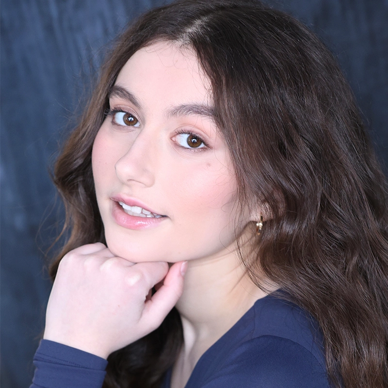 A young woman with wavy brown hair and fair skin poses against a dark background. She wears a navy blue top and rests her chin on her hand, looking at the camera with a gentle, confident expression—evoking the poise of Sawyer Whitted.