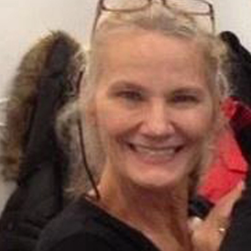 Candace Tovar, a smiling older woman with light skin and long blond hair, wears glasses atop her head. She is indoors in dark clothing, standing before a wall with coats hanging—including a dark coat with a fur-lined hood.