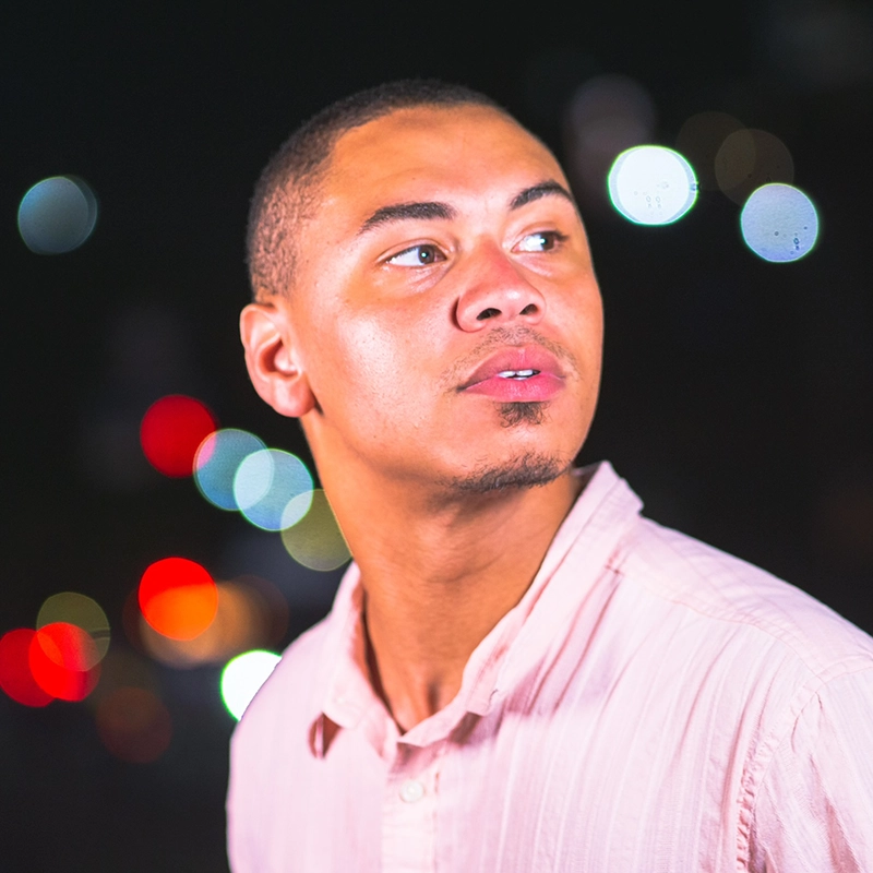 A young man with short hair and a trimmed goatee wears a pale pink, collared shirt and looks slightly to the left. The background is dark, with colorful, out-of-focus lights creating a bokeh effect.