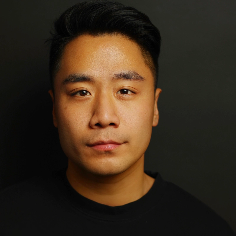 A young man with short black hair and smooth skin is facing the camera against a plain dark background. He has a neutral expression and is wearing a black crew-neck shirt. The lighting is even, highlighting his facial features clearly.