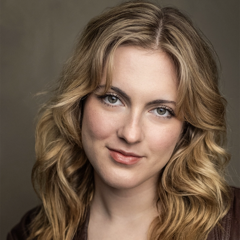 A woman with wavy, shoulder-length blonde hair and light eyes looks directly at the camera. She has a fair complexion, subtle makeup, and a slight smile. The background is plain and softly lit, highlighting her facial features.