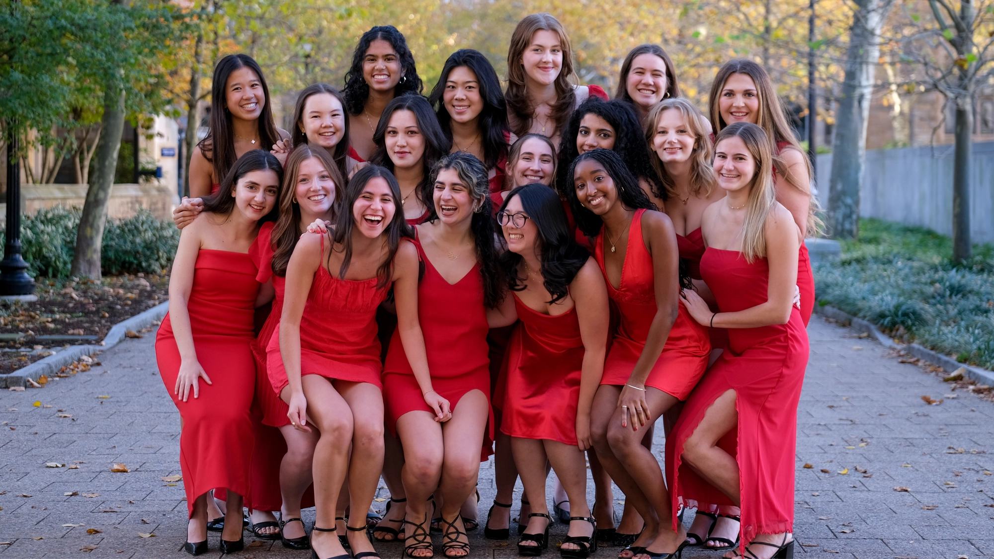 A group of eighteen young women from Yale University’s Proof of the Pudding pose together outdoors on a walkway lined with trees. They are all smiling and wearing red dresses, with autumn leaves and a stone wall in the background.