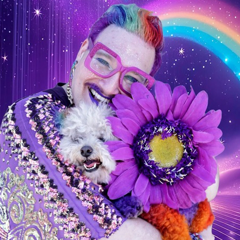 A joyful person with rainbow hair, purple lipstick, and oversized pink glasses hugs a fluffy white dog. Both wear vibrant purple and gold costumes. A large purple flower covers part of the dog. Artist Zach Aaronson creates a starry, rainbow-filled scene.
