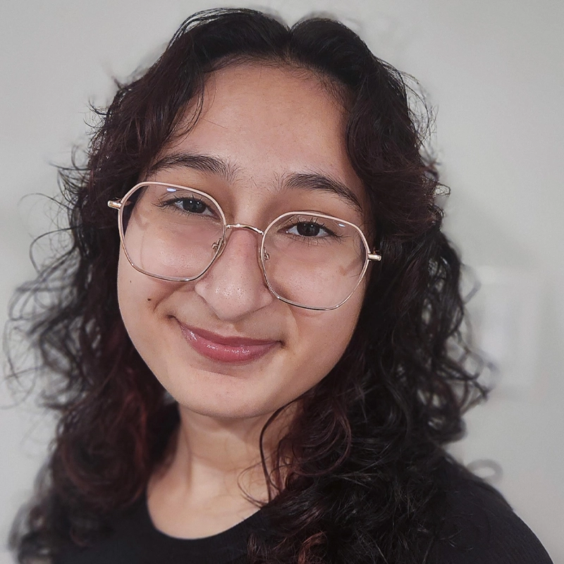 Olivie Aiere smiles softly, her wavy dark brown hair and light brown skin framed by large round glasses with thin, gold rims. Dressed in a black top, she stands against a neutral, softly blurred background that draws focus to her face.