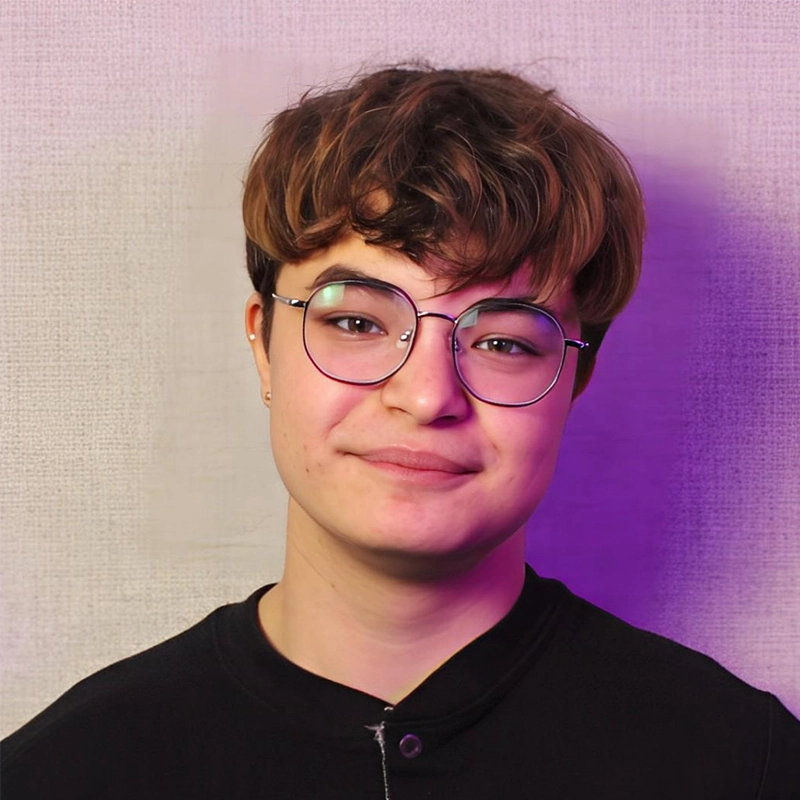 A young person with short, wavy brown hair and round glasses smiles softly at the camera. They wear a black top and stand against a textured light gray background with a purple light cast on the right side.