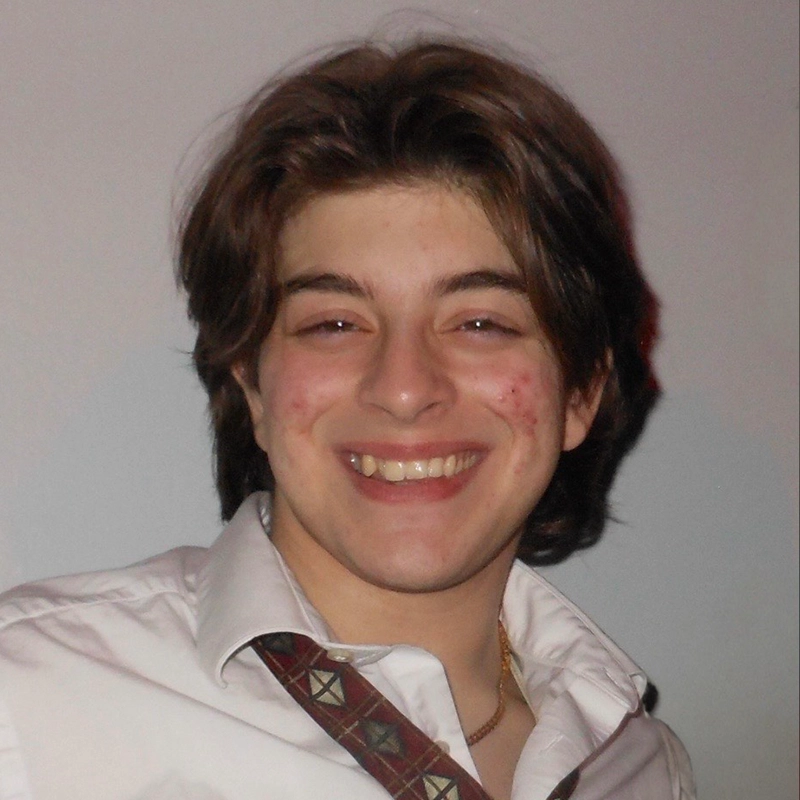 A young person with medium-length brown hair and acne smiles at the camera. They wear a white collared shirt, partially unbuttoned, with a patterned tie draped loosely around their neck. The background is a plain, light-colored wall.