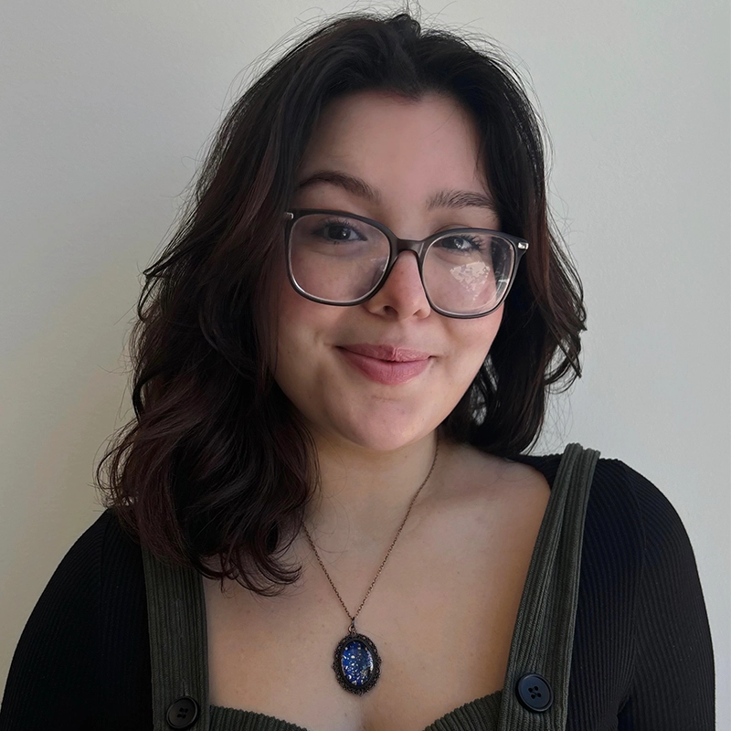 Amanda Negrete, a young woman with wavy dark hair and glasses, smiles softly. She wears a black top with wide straps and large buttons, along with a necklace featuring a blue pendant. The background is a plain, light-colored wall.