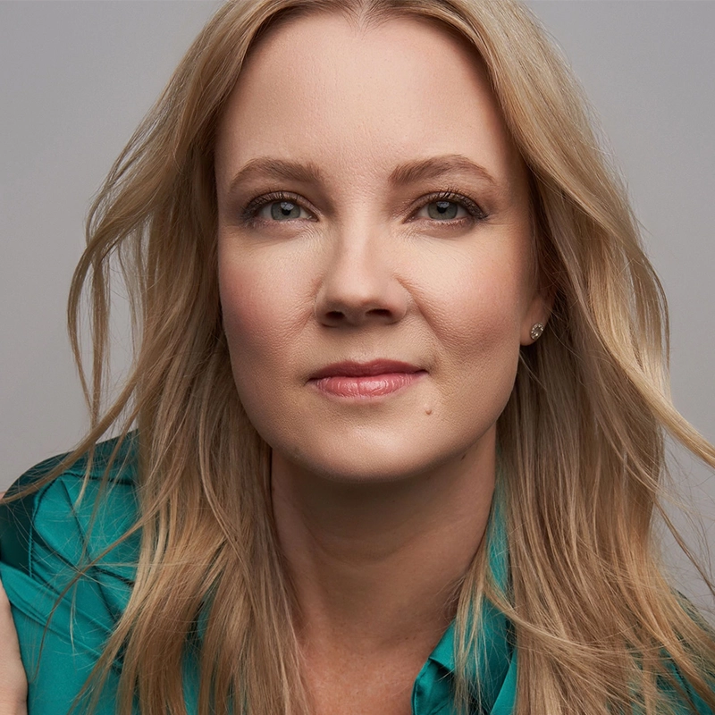 A woman with fair skin and long blonde hair looks at the camera with a slight smile. She is wearing a teal blouse and small stud earrings. The softly tousled hair frames her face, captured in the signature style of Ryan Williams against a light gray background.