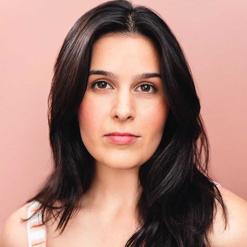 A woman with long, dark brown hair and fair skin looks directly at the camera with a neutral expression. She is wearing a sleeveless top and stands against a soft pink background. Her makeup is natural with subtle blush on her cheeks.