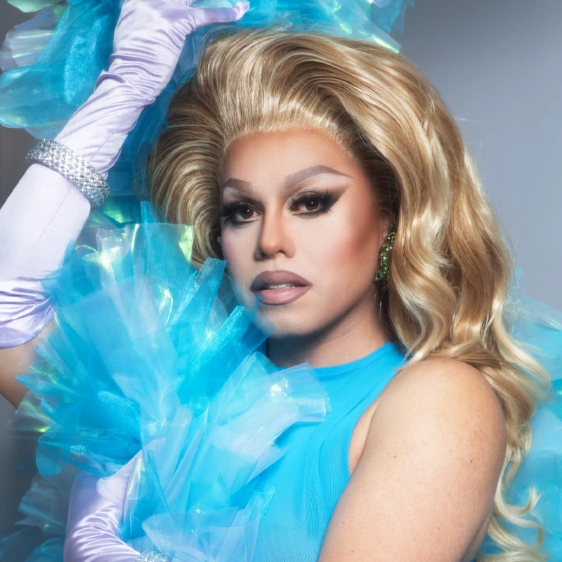A glamorously dressed drag queen, Pixie Aventura, with voluminous blond hair poses in a blue sheer outfit. She wears dramatic makeup, sparkling earrings, long lavender gloves, and holds blue tulle, exuding confidence and elegance.