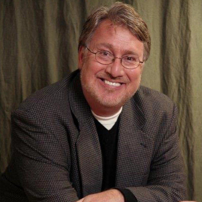 A middle-aged man with gray hair, glasses, and a beard smiles warmly at the camera. He is wearing a checkered sport coat over a black sweater and white shirt. The background is a greenish curtain. His hands are resting in front of him.
