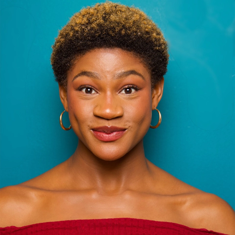 Aeja Barrows, a young woman with short curly hair dyed brown at the top, smiles slightly in gold hoop earrings and a red off-shoulder top. She stands against a bright teal background, exuding confidence and friendliness.