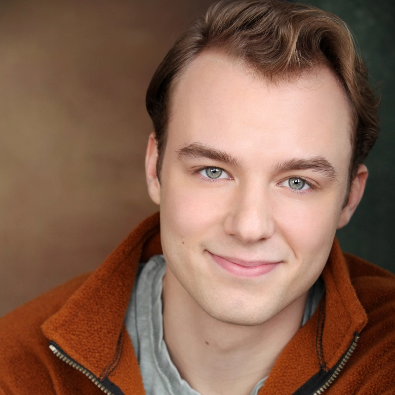 A young man with light skin, blue eyes, and short, light brown hair smiles softly at the camera. Henry Beddoe wears a rust-colored fleece jacket over a gray shirt, with a softly blurred background in warm brown and green tones.