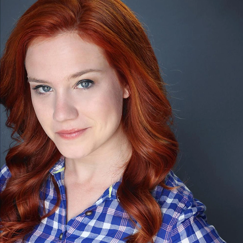 Hayden DeWitt, a woman with long, wavy red hair and fair skin, smiles softly. She wears a blue and white checkered shirt and poses before a plain dark gray background, looking directly at the camera with a calm expression.