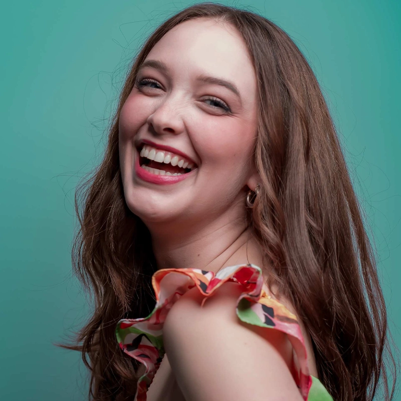 A young woman with long brown hair smiles brightly, showing her teeth. She wears a colorful, ruffled sleeveless top and has a light complexion. The background is a solid teal color, creating a cheerful and vibrant portrait.