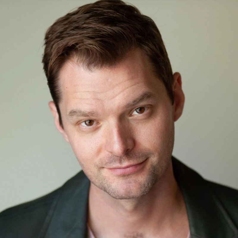 Ryan Williams, a man with short brown hair and light skin, wears a dark jacket over a light shirt. He looks slightly upward with a slight smile against a plain, neutral background. Soft lighting highlights his facial features.